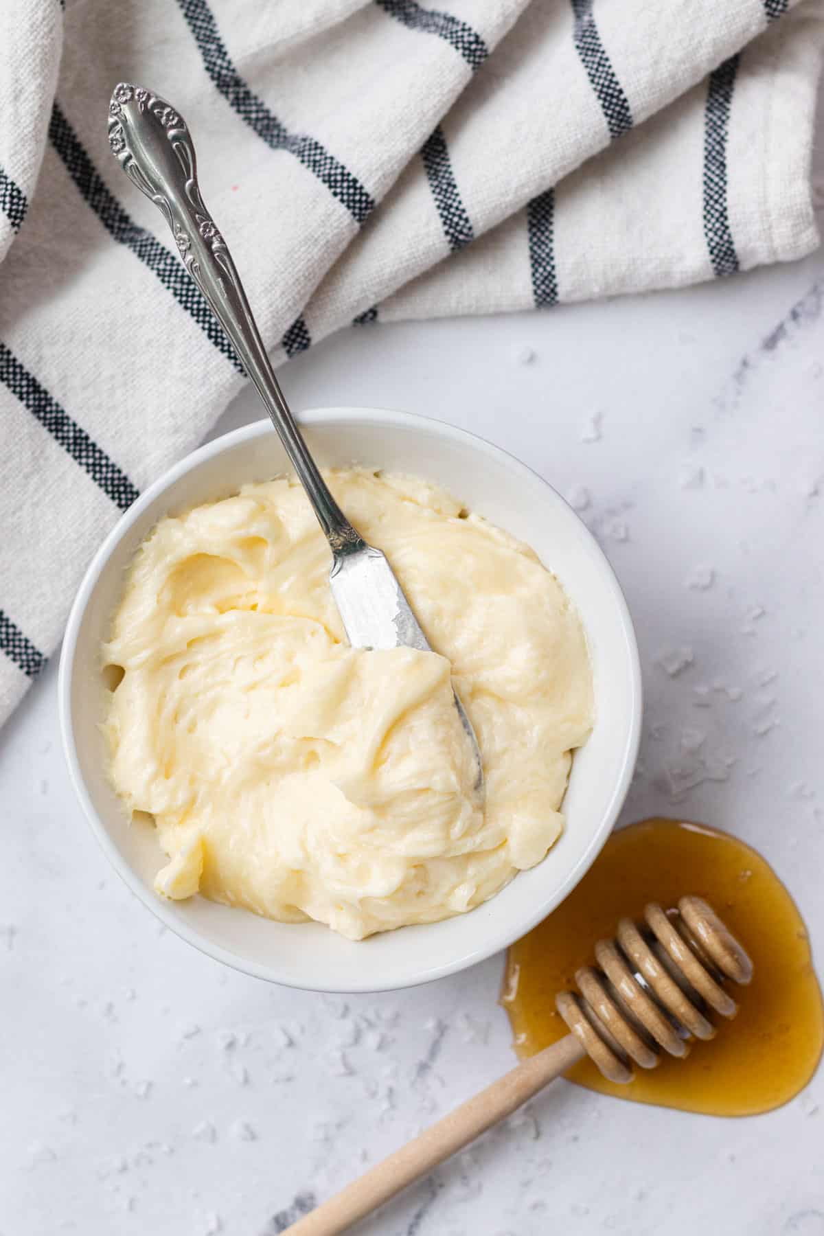 A white bowl filled with creamy whipped honey butter with a metal butter knife resting inside, placed next to a wooden honey dipper in a small puddle of honey.