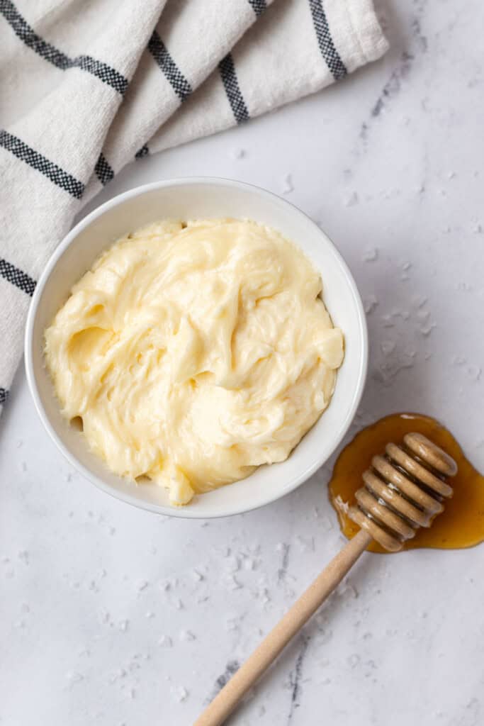 A white bowl of whipped honey butter next to a wooden honey dipper in a puddle of honey.