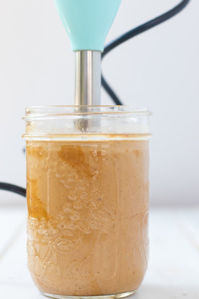 Blending peanut sauce in a clear jar with a blue immersion blender.