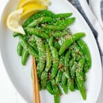 A large white serving plate filled with garlic sesame snow peas, a pair of chopsticks, and wedges of lemon on the side.