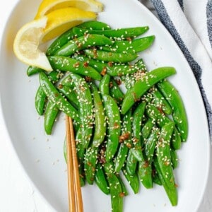 A large white serving plate filled with garlic sesame snow peas, a pair of chopsticks, and wedges of lemon on the side.