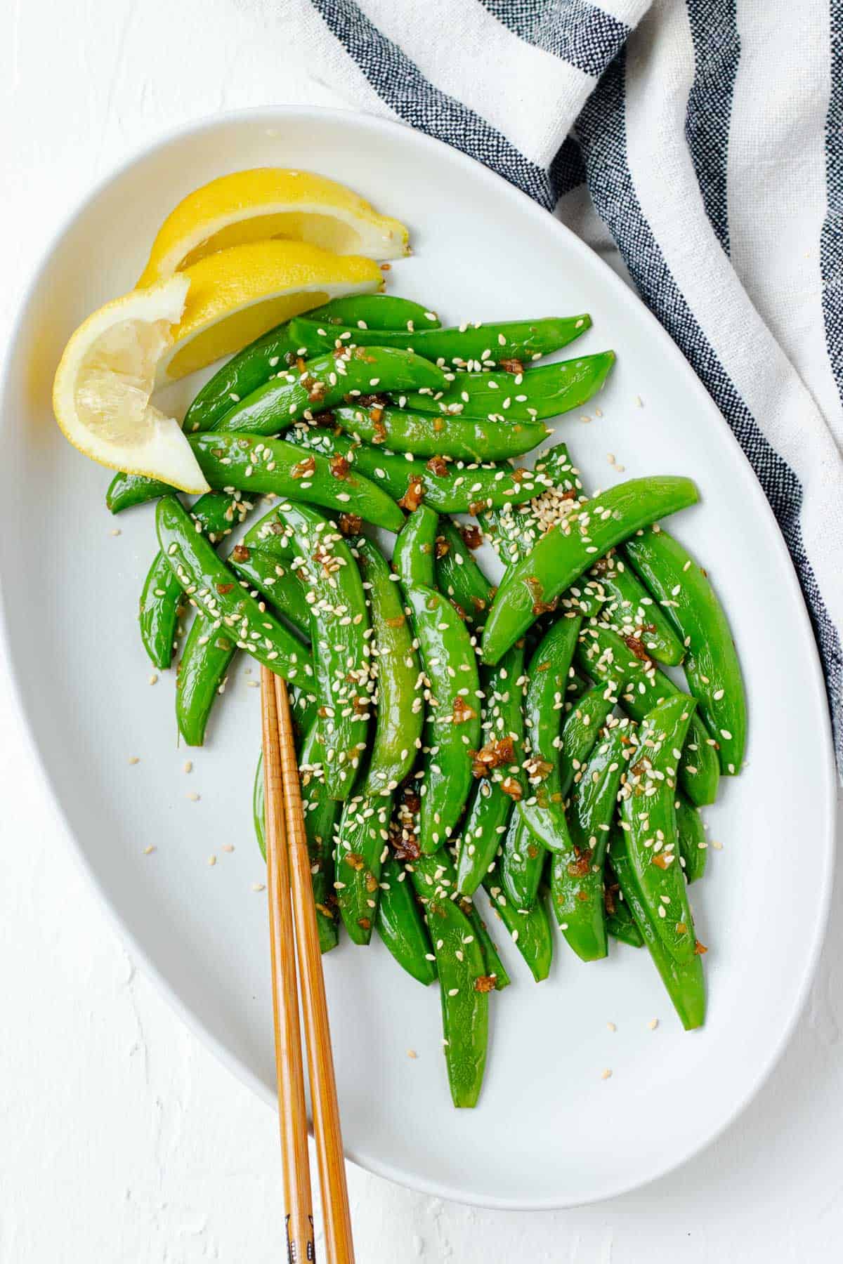A large white serving plate filled with garlic sesame snow peas, a pair of chopsticks, and wedges of lemon on the side.