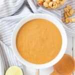 A white bowl full of Vietnamese peanut sauce with a bowl of peanuts, half a lime, and a wooden spoon on the side.
