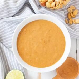 A white bowl full of Vietnamese peanut sauce with a bowl of peanuts, half a lime, and a wooden spoon on the side.