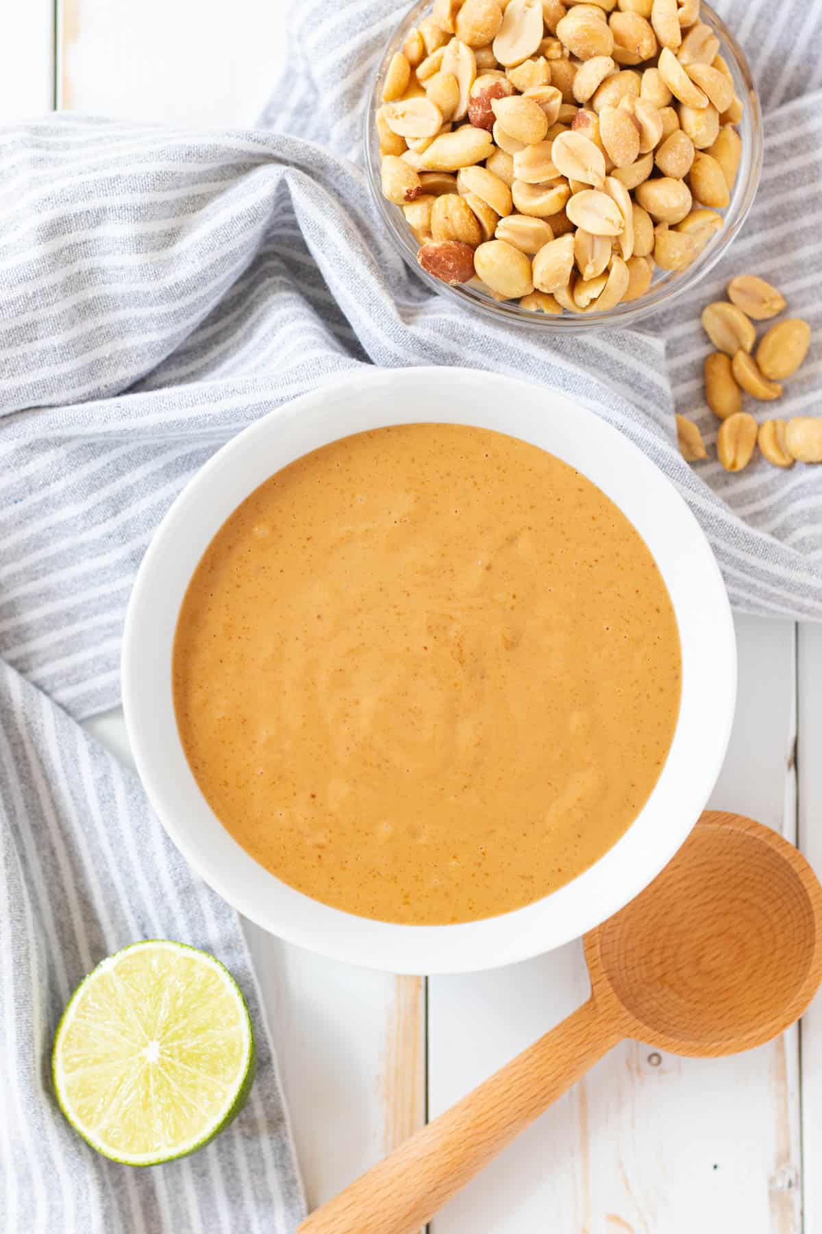 A white bowl full of Vietnamese peanut sauce with a bowl of peanuts, half a lime, and a wooden spoon on the side.