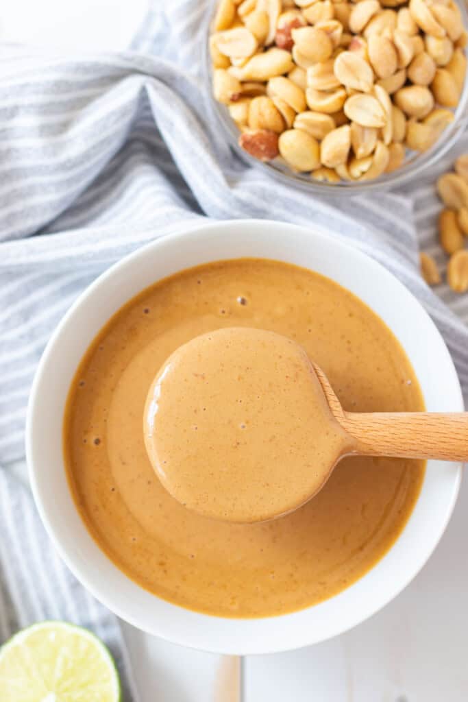 A wooden spoon hovering a scoop of creamy peanut sauce over a white bowl full.