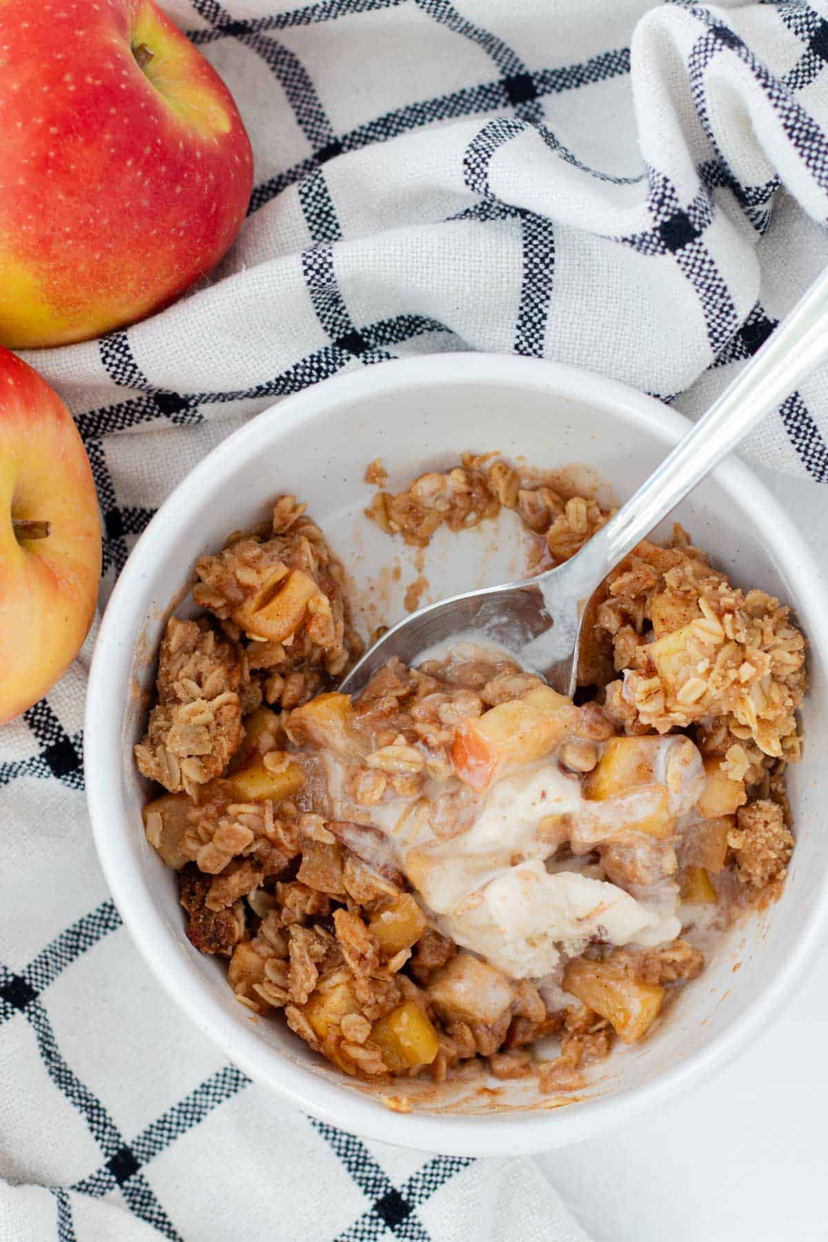 A bowl of apple crisp half eaten with a spoon and melted ice cream starting to mix in.