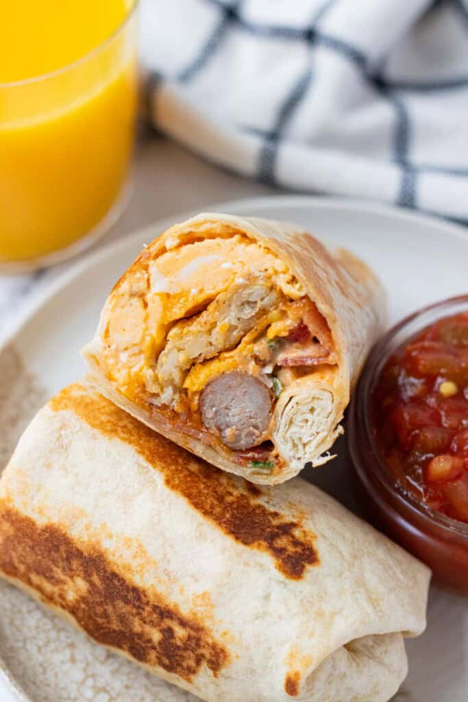 Fully loaded breakfast burrito cut in half on a plate with a side of salsa and orange juice.