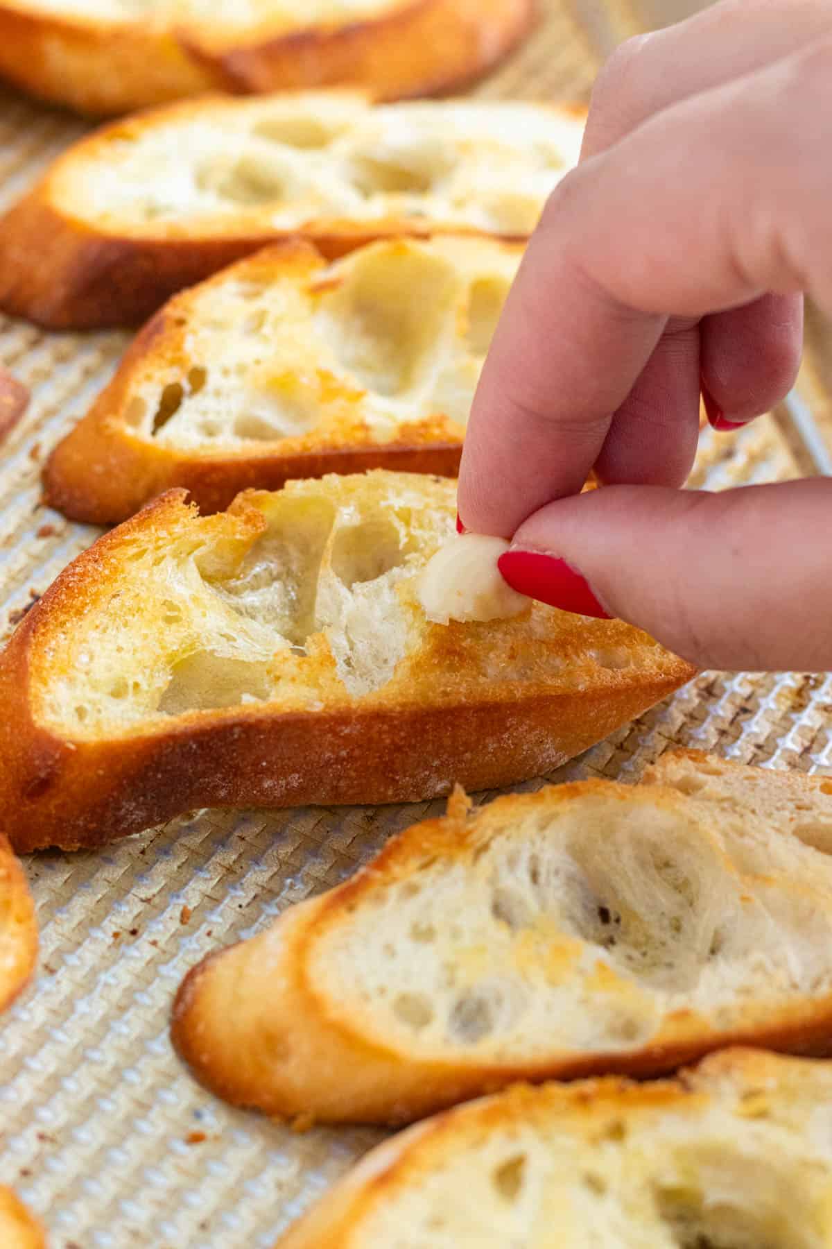 A hand holding a garlic clove, rubbing it on the toasted baguette slices.