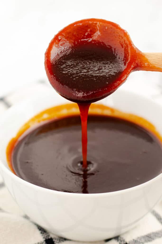 Thick brown barbecue sauce drizzling from a wooden spoon into a bowl, showing the consistency of the finished sauce.