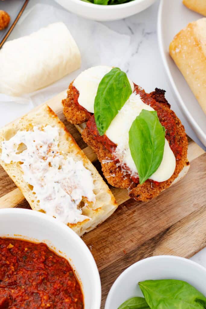 Building the chicken parm sub by spreading roasted garlic aioli on the baguette and topping with chicken parmesan cutlet and fresh basil.