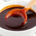 Thick, glossy brown sauce in a white bowl with a wooden spoon lifting a spoonful to show its smooth consistency.