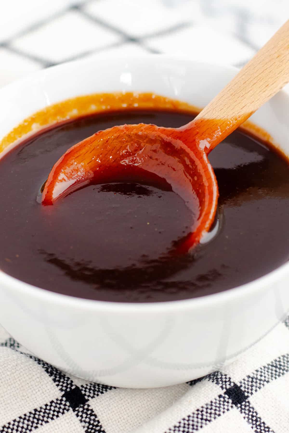 Thick, glossy brown sauce in a white bowl with a wooden spoon lifting a spoonful to show its smooth consistency.