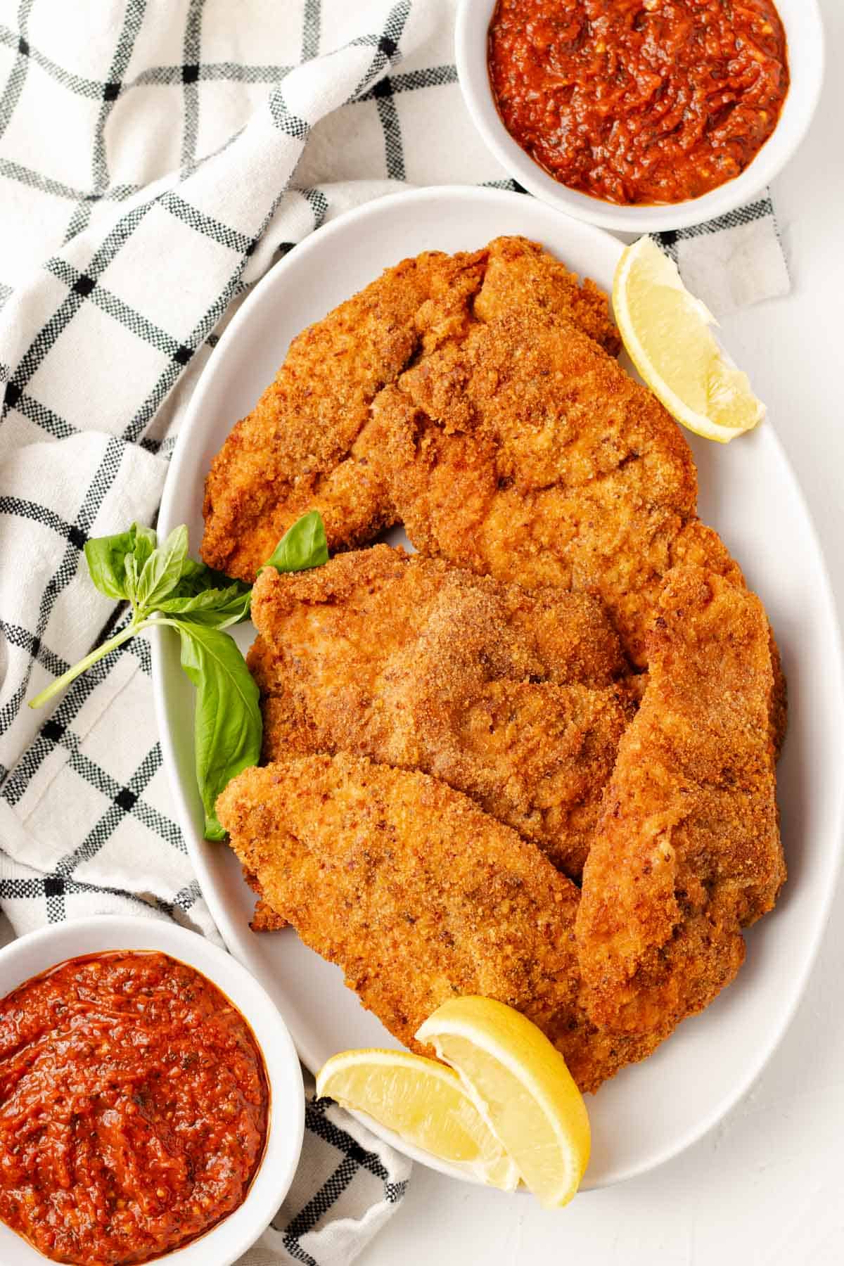 Golden crispy chicken cutlets arranged on a plate with lemon wedges, fresh basil, and marinara dipping sauce.