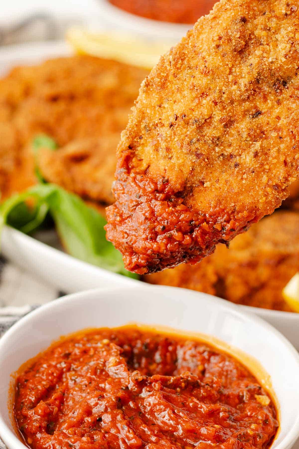 Crispy breaded chicken cutlet dipped into marinara sauce with more chicken cutlets in the background.