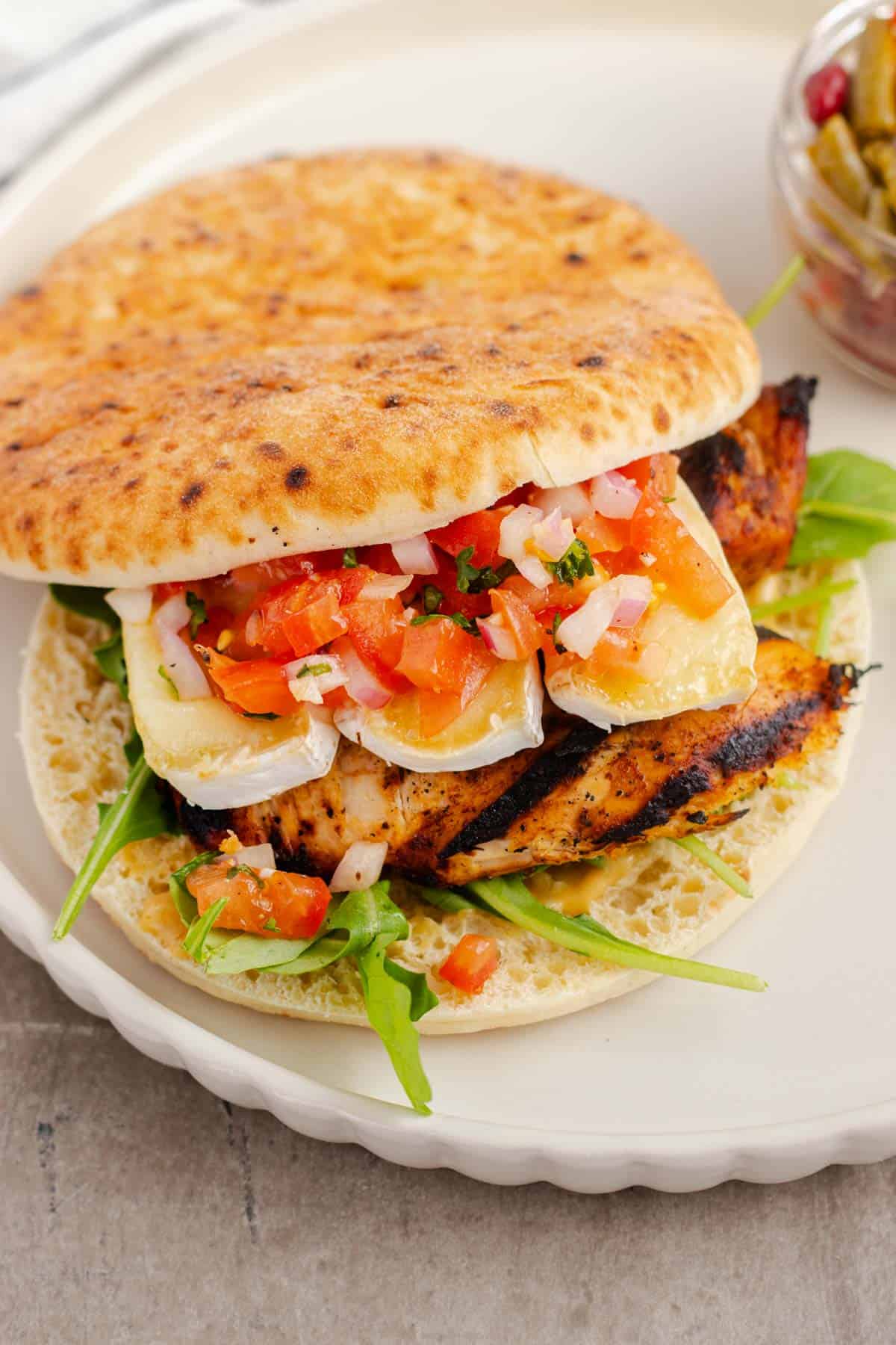 Grilled chicken sandwich with melted brie, arugula, and fresh tomato bruschetta on a white scalloped-edge plate.