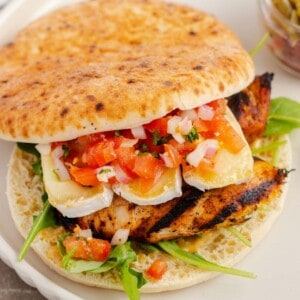 Grilled chicken sandwich with melted brie, arugula, and fresh tomato bruschetta on a white scalloped-edge plate.