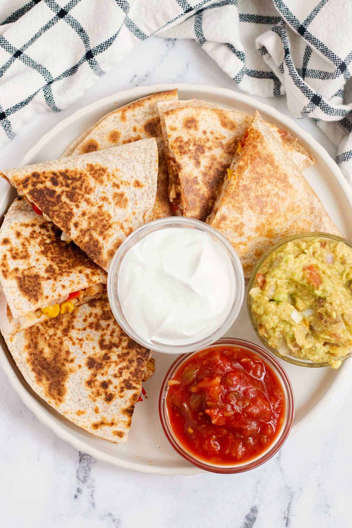 Cooked quesadilla triangles on a white plate with guacamole, sour cream, and salsa.