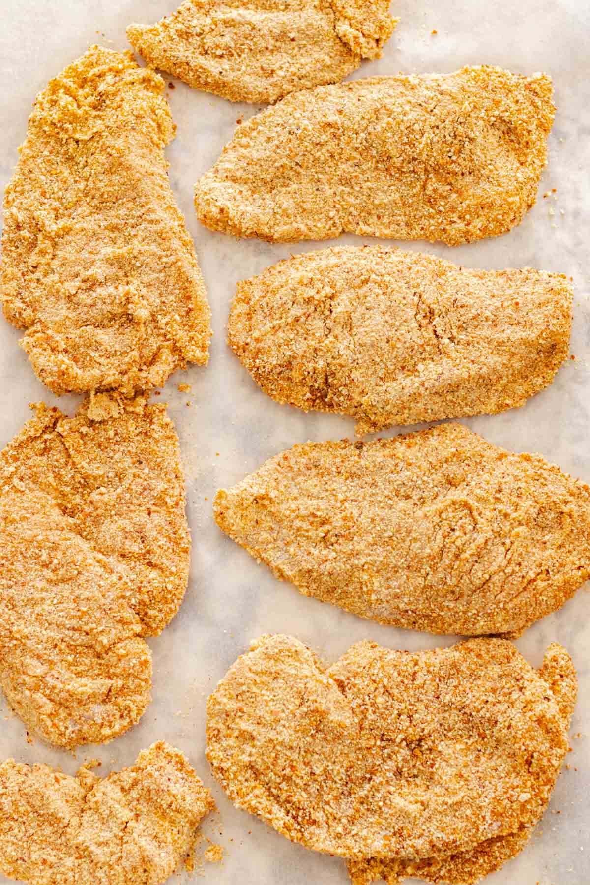 Raw chicken cutlets, breaded on parchment paper.