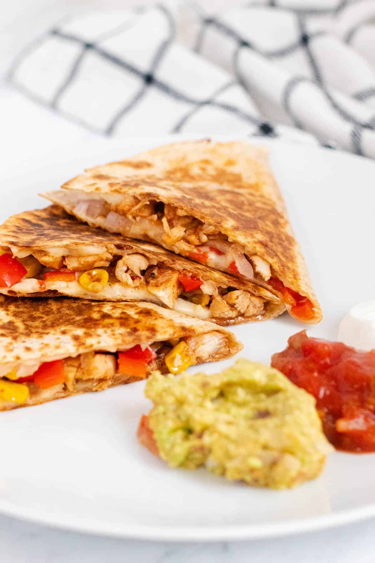 BBQ chicken quesadilla triangles cut on a white plate with salsa, guacamole, and sour cream dipping sauces on the side.