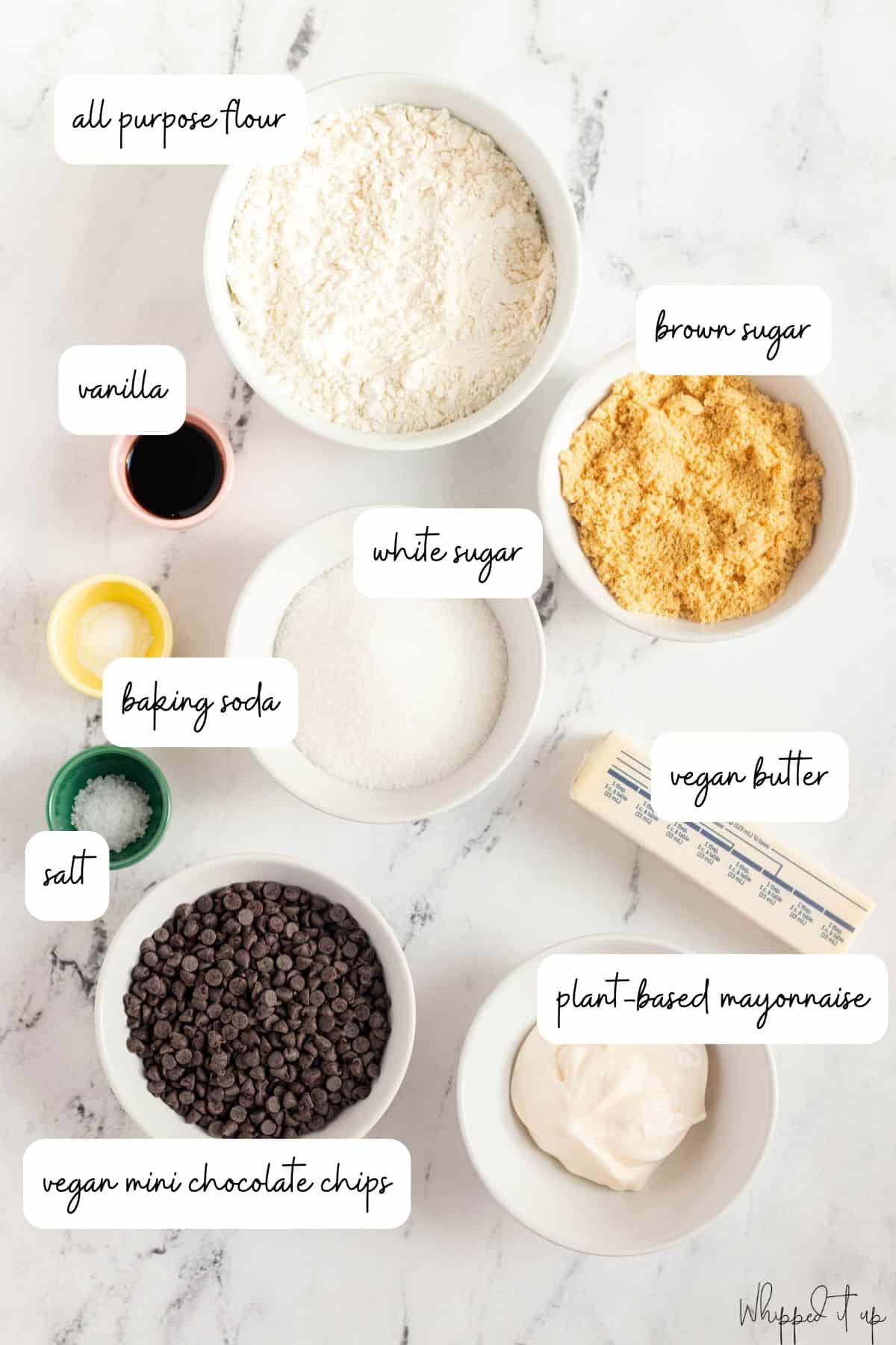 Ingredients to make cookie dough are measured out in white bowls on a marble background.