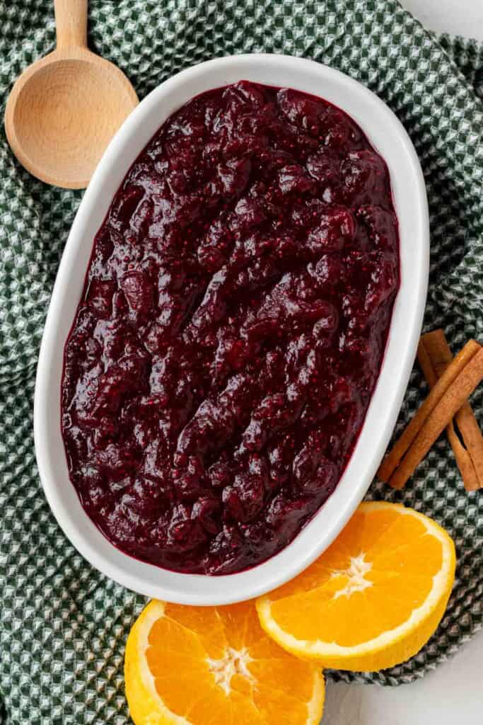 A white serving dish full of whole berry cranberry sauce with an orange and cinnamon sticks on the side.